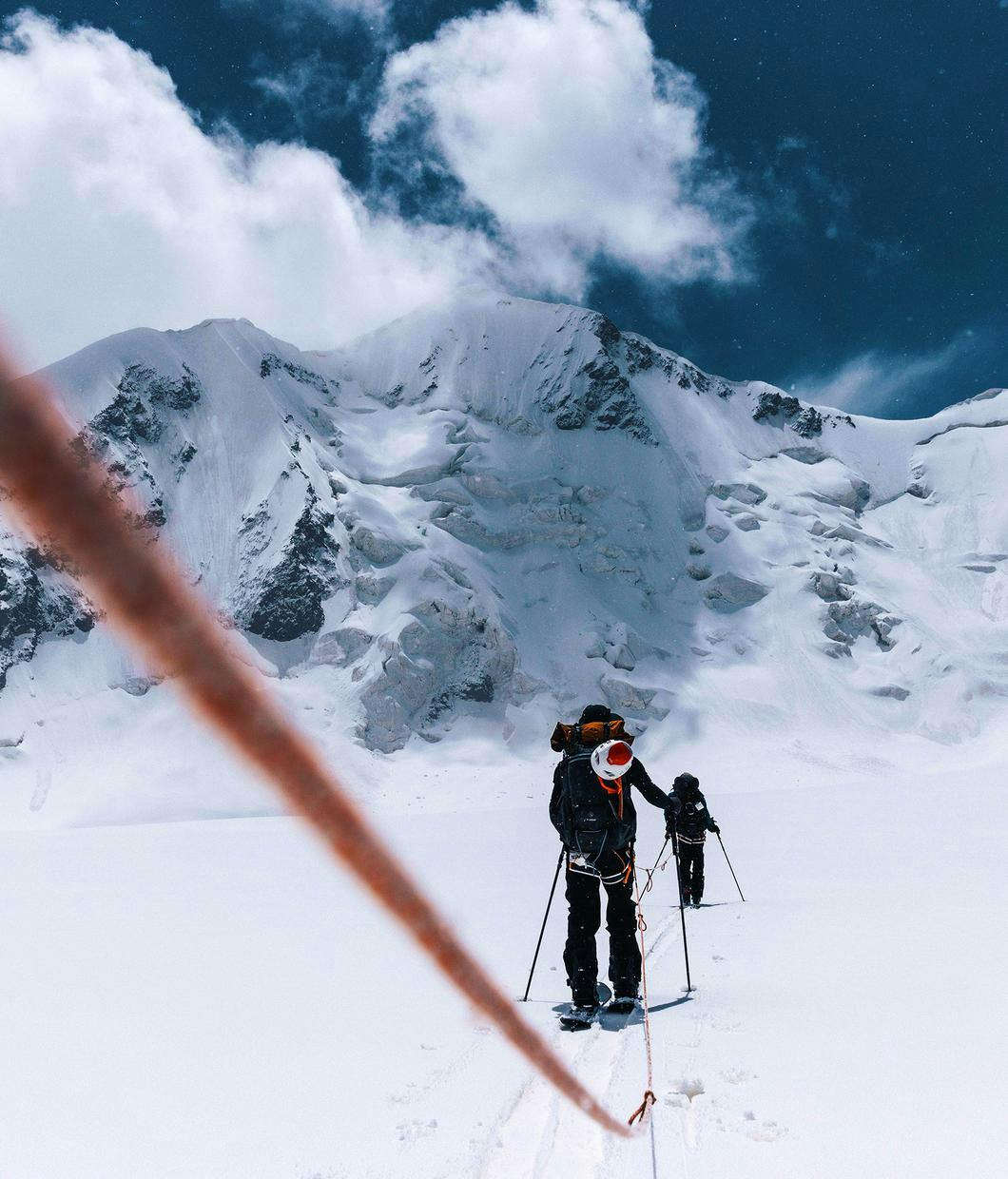 3 Bergsteiger gehen an einem Seil hintereinander auf einer Ebene aus Schnee in Richtung eines Gipfel. Dieser ist im Hintergrund zusehen. Ein weißer Spitziger breiter Gipfel, dahinter blauer Himmel und eine weiße Wolke im Bereich des Gipfel. © ┬®_Jeremy Bernard